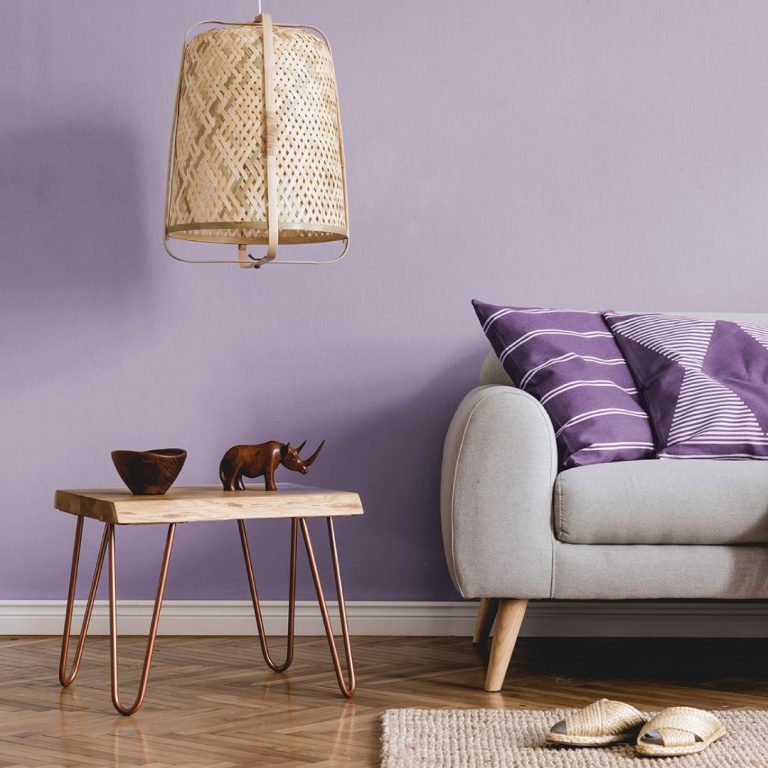 Living in stil minimalist - perete lila, canapea cu 2 perne alb-mov in dungi, rinocer din lemn, bol din lemn, masuta cu 4 picioare bronz, covor, papuci